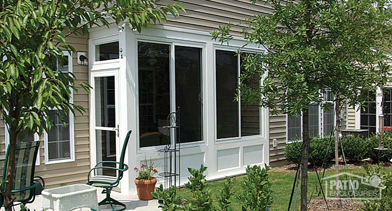 A white sunroom patio enclosure with solid knee walls and trees and shrubs all around.
