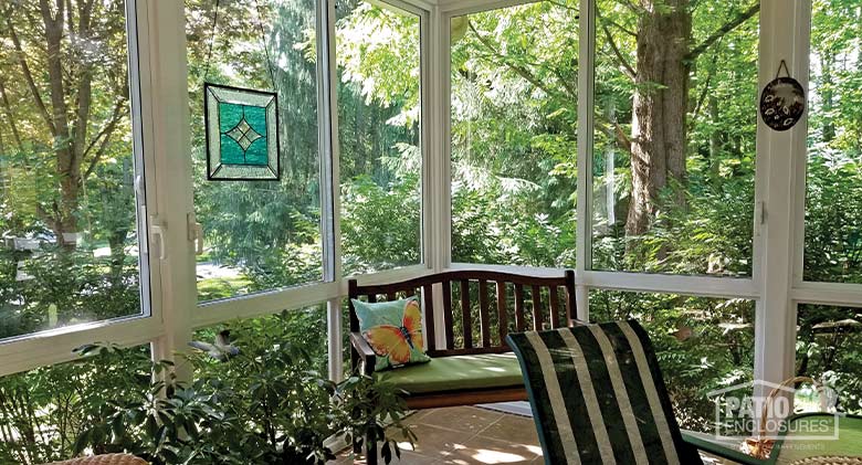 A shady and cool-looking sunroom surrounded by trees and bushes furnished with a bench & butterfly pillow and stained glass.