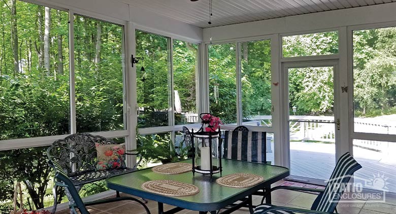A glass dining table and chairs with striped cushions inside a glass sunroom surrounded by trees and bushes.

