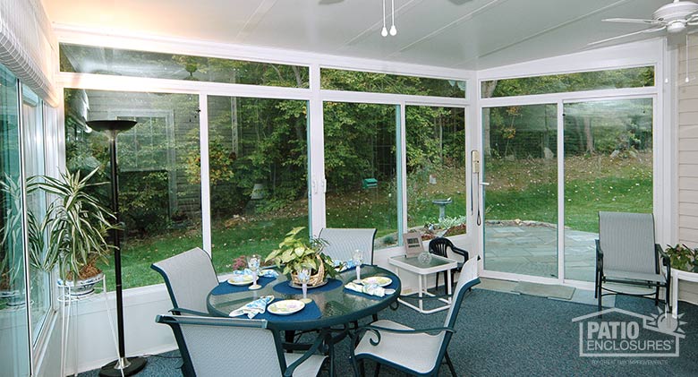 A glass table and chairs set for dinner inside a glass sunroom with solid knee walls, transom, and blue carpet.