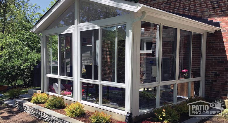 The exterior of a white sunroom with gable roof, glass wings and glass knee walls. Neat plantings around the room.