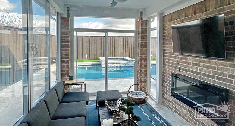 Interior of a patio enclosure with seating area facing a mounted TV and electric fireplace with a view of an in-ground pool.