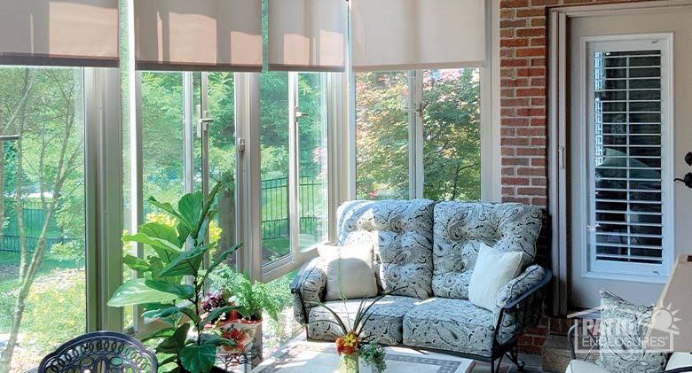 Comfortable furniture and potted plants in a glass patio enclosure with sun streaming in below partially closed roller shades.