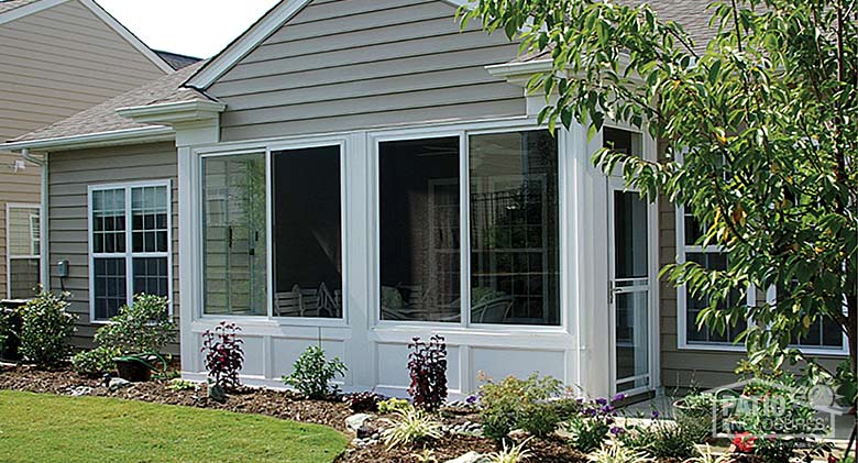 The exterior of an L-shaped patio enclosure with white frame and metal roof situated behind a large in-ground pool.