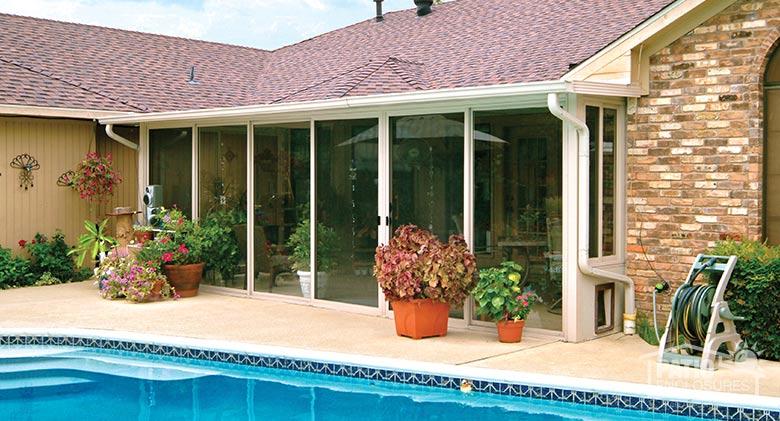 Glass sunroom patio enclosure with dog door overlooking a lovely pool and bright potted plants and flowers on the patio.