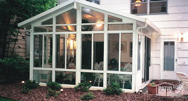 A gabled-roof white sunroom with glass wings, transoms, and a glass knee wall with small bushes in front.