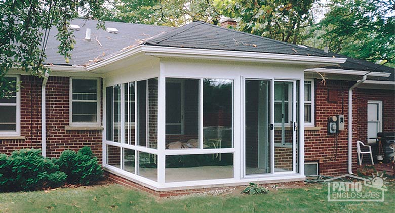  A glass sunroom with glass knee wall, sliding doors and hip roof on the concrete patio of a brick home.