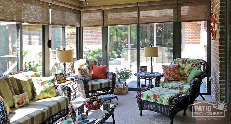 Brown wicker furniture with bright cushions and pillows in a brown glass sunroom with open roller shades in a lighter brown.