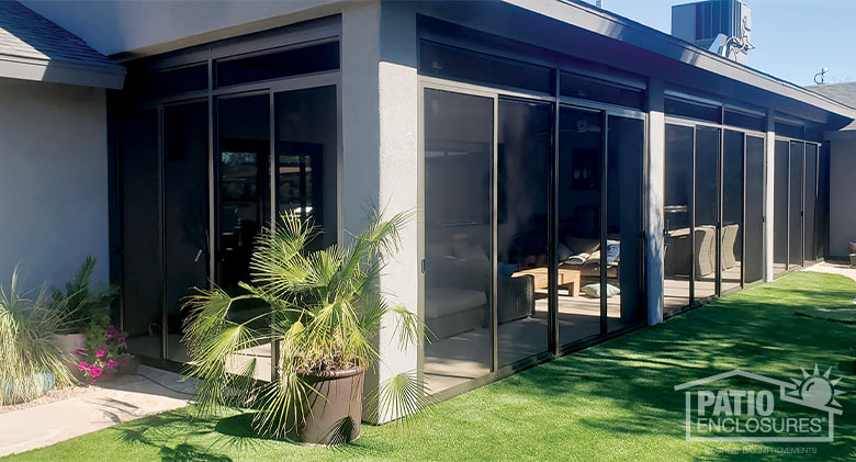 Exterior of a covered patio enclosed with brown-framed screens and gray stucco pillars for support. Potted plants outside.