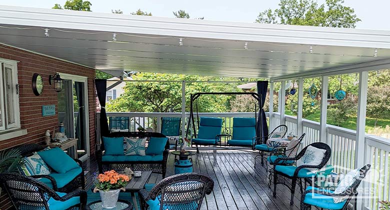 A white roof over most of a large deck with comfortable chairs, benches and a swing with pillows and blue cushions.