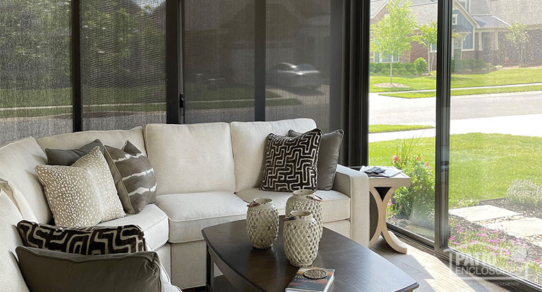A front porch or lanai enclosed in glass with a white sofa, brown patterned pillows, and brown window shades.