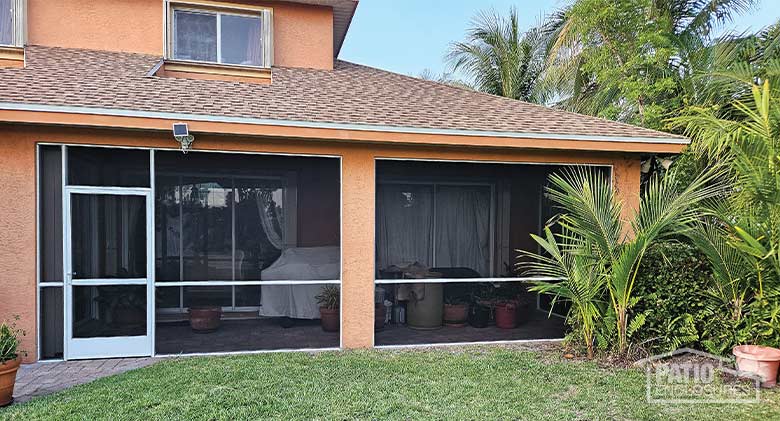 A peach stucco home with a white-framed lanai enclosure with a storm door. Tropical plants outside the room.