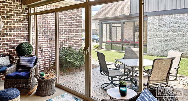 A glass lanai enclosure with blue striped cushions on wicker chairs inside, a glass table, and beige chairs outside.